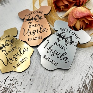 Op de afbeelding: Drie babymeisjes welkom-versieringen in goud, zilver en ros&eacute; goud. Elke versiering is in de vorm van een rompertje en heeft een strik, de tekst "Welkom", "Baby Ursula" en de datum "8.21.2021".