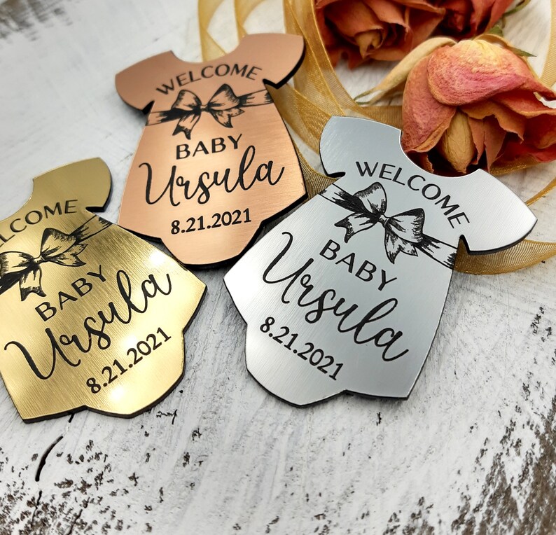 Op de afbeelding: Drie babymeisjes welkom-versieringen in goud, zilver en ros&eacute; goud. Elke versiering is in de vorm van een rompertje en heeft een strik, de tekst "Welkom", "Baby Ursula" en de datum "8.21.2021".