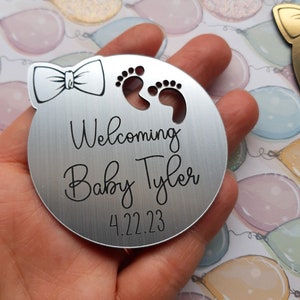 Op de afbeelding: Zilveren rond ornament met een strik, voetafdrukken en de tekst "Welkom Baby Tyler 4.22.23".