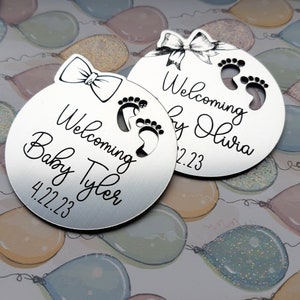 Op de afbeelding: Twee zilveren ronde ornamenten met een strik en babyvoetjes. De tekst op de ornamenten luidt "Welkom Baby Tyler 4.22.23" en "Welkom Baby Olivia 2.23".