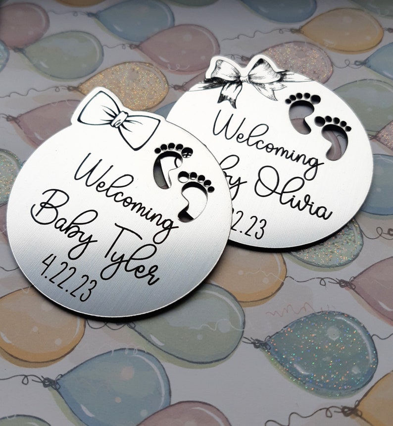Op de afbeelding: Twee zilveren ronde ornamenten met een strik en babyvoetjes. De tekst op de ornamenten luidt "Welkom Baby Tyler 4.22.23" en "Welkom Baby Olivia 2.23".