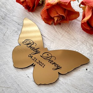 May include: Gold butterfly-shaped metal ornament with the text "Baby Darcy 04.21.2021" engraved on it.