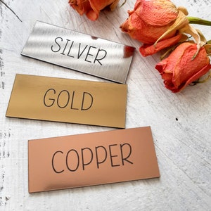 Op de afbeelding: Drie rechthoekige metalen labels met de woorden "SILVER", "GOLD" en "COPPER" erop gegraveerd. De labels zijn verticaal op een wit houten oppervlak gerangschikt.