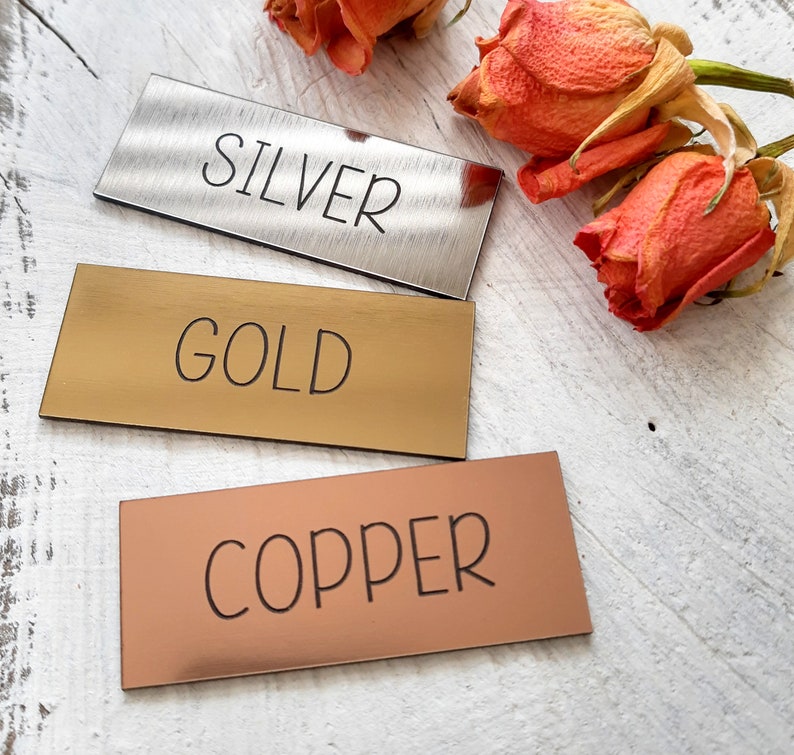 Op de afbeelding: Drie rechthoekige metalen labels met de woorden "SILVER", "GOLD" en "COPPER" erop gegraveerd. De labels zijn verticaal op een wit houten oppervlak gerangschikt.