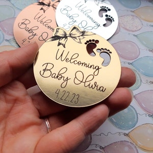 Op de afbeelding: Rond gouden ornament met een strik en voetafdruk-uitsnijdingen. Het ornament zegt "Welkom Baby Olivia 4.22.23".