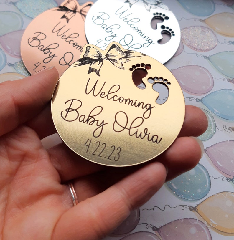 Op de afbeelding: Rond gouden ornament met een strik en voetafdruk-uitsnijdingen. Het ornament zegt "Welkom Baby Olivia 4.22.23".