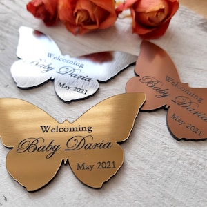 Może przedstawiać: Trzy złote, srebrne i miedziane metalowe ozdoby w kształcie motyla z wygrawerowanym tekstem "Welcoming Baby Daria May 2021" na każdym.