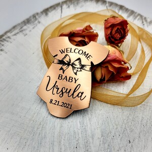 Op de afbeelding: Koperkleurig babymeisjes ornament in de vorm van een jurk met de tekst "Welcome Baby Ursula 8.21.2021" erop gegraveerd.