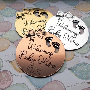 Op de afbeelding: Drie ronde, gegraveerde, metalen babyshower-cadeautjes in goud, zilver en koper. Elk cadeautje heeft een strik, voetafdrukken en de tekst "Welkom Baby Olivia" met de datum "4.22.23" op het koperen cadeautje.