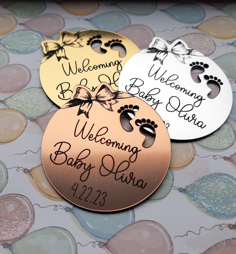 Op de afbeelding: Drie ronde, gegraveerde, metalen babyshower-cadeautjes in goud, zilver en koper. Elk cadeautje heeft een strik, voetafdrukken en de tekst "Welkom Baby Olivia" met de datum "4.22.23" op het koperen cadeautje.