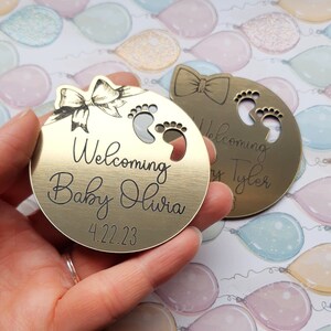 Op de afbeelding: Twee ronde metalen ornamenten in goudkleur met gegraveerde tekst. De eerste ornament heeft een strik en de tekst "Welcoming Baby Olivia 4.22.23". De tweede ornament heeft een strik en de tekst "Welcoming Baby Tyler".