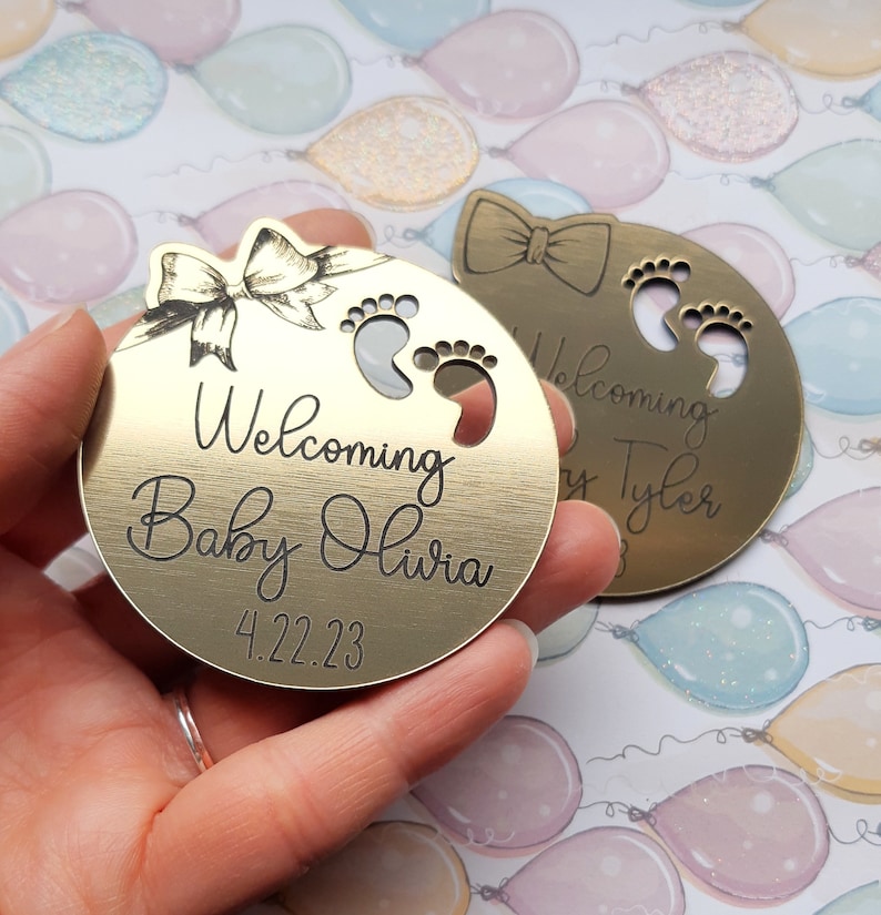 Op de afbeelding: Twee ronde metalen ornamenten in goudkleur met gegraveerde tekst. De eerste ornament heeft een strik en de tekst "Welcoming Baby Olivia 4.22.23". De tweede ornament heeft een strik en de tekst "Welcoming Baby Tyler".