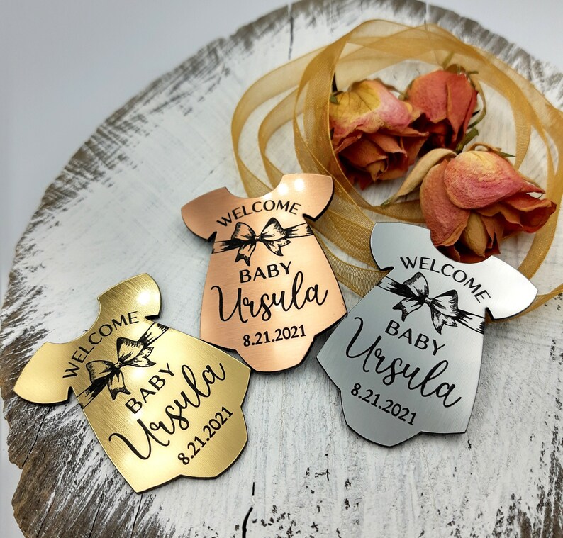 Op de afbeelding: Drie babyshower-cadeautjes in de vorm van een jurk met de tekst "Welkom Baby Ursula 8.21.2021". De cadeautjes zijn gemaakt van goud, zilver en ros&eacute; goud.