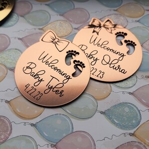 Op de afbeelding: Twee ronde baby-aankondigingsetiketten van koperkleurig metaal met een strik en voetafdrukken. De tekst op de etiketten luidt "Welkom Baby Tyler 4.22.23" en "Welkom Baby Olivia 4.22.23".