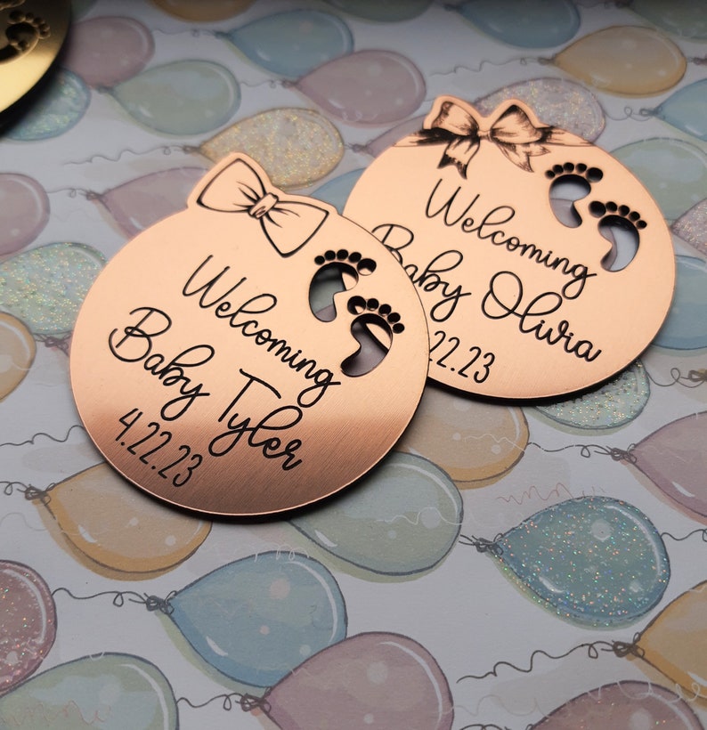 Op de afbeelding: Twee ronde baby-aankondigingsetiketten van koperkleurig metaal met een strik en voetafdrukken. De tekst op de etiketten luidt "Welkom Baby Tyler 4.22.23" en "Welkom Baby Olivia 4.22.23".