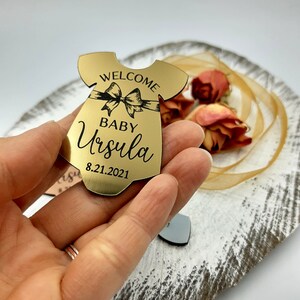 Op de afbeelding: Gouden baby-rompertje-vormige ornament met de tekst "Welcome Baby Ursula 8.21.2021" erop gegraveerd.