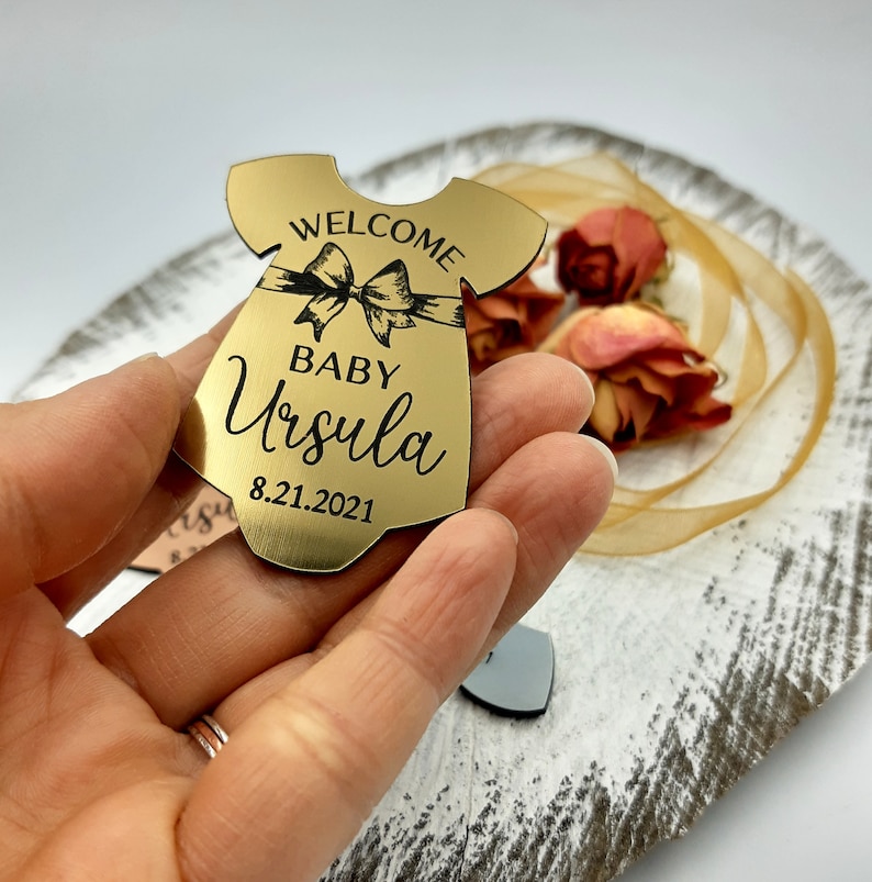 Op de afbeelding: Gouden baby-rompertje-vormige ornament met de tekst "Welcome Baby Ursula 8.21.2021" erop gegraveerd.