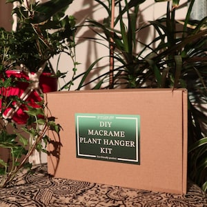 May include: A brown cardboard box with a green and white label that says "DIY Macrame Plant Hanger Kit" and "Eco-friendly product". The box is sitting on a patterned fabric surface.