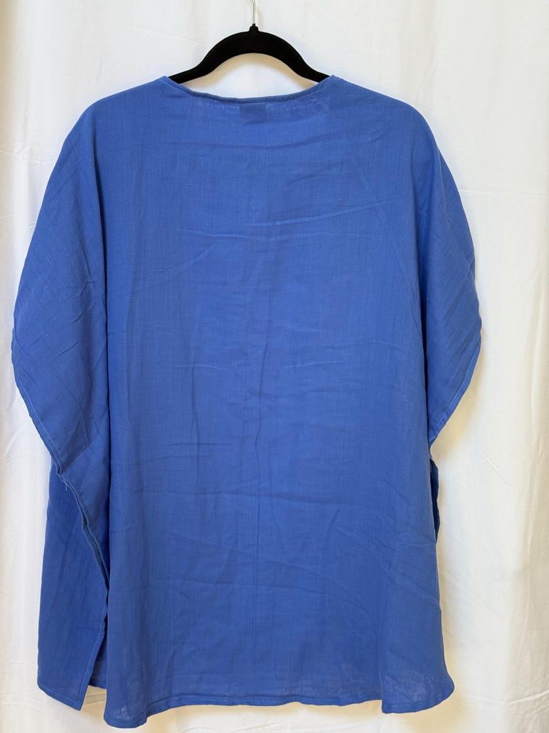 May include: A solid cornflower blue tunic top with a round neckline and short, wide sleeves. The garment features a loose, relaxed fit with side slits at the hem, constructed from a lightweight, opaque fabric that hangs straight down from the shoulders.
