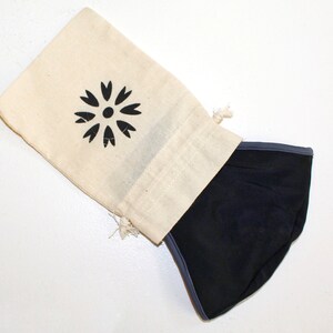 May include: A black face mask with a grey trim is folded and placed inside a white cotton drawstring bag. The bag has a black floral design printed on it.
