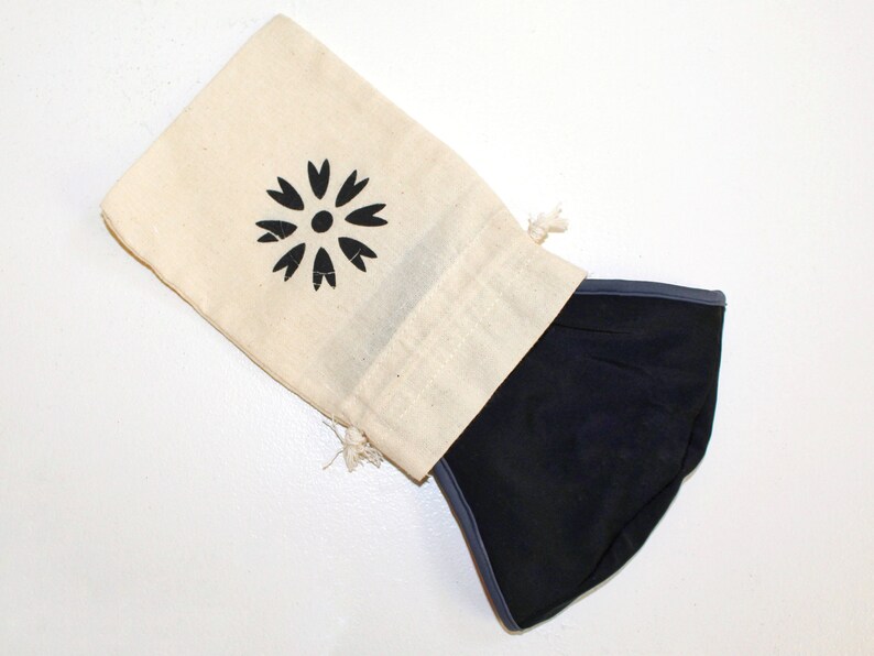 May include: A black face mask with a grey trim is folded and placed inside a white cotton drawstring bag. The bag has a black floral design printed on it.