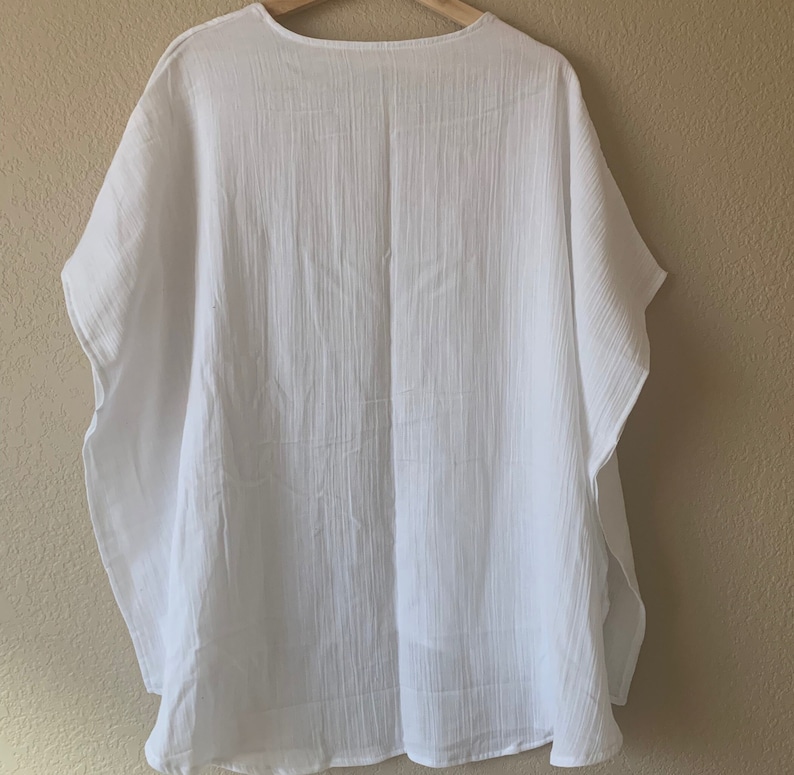 May include: A solid white kaftan top featuring a lightweight, crinkled fabric texture with a loose, flowing silhouette, short wide sleeves, and a simple rounded neckline, presented on a wooden hanger against a neutral wall background.