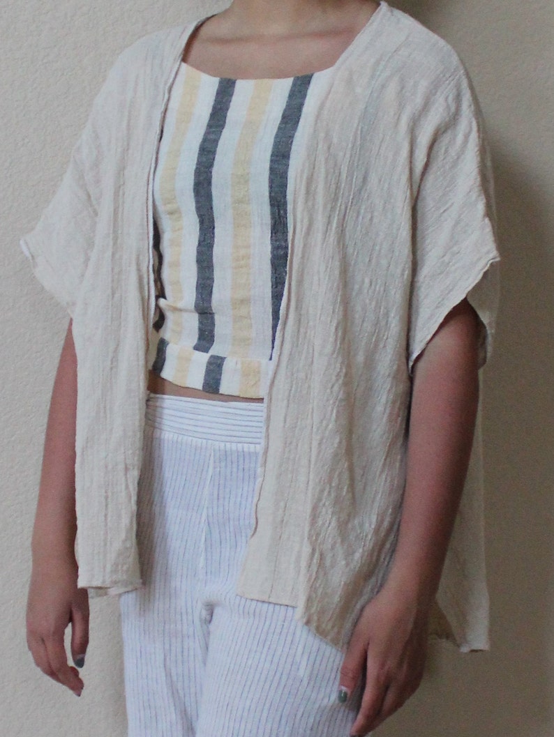 May include: A beige open-front kimono-style cardigan with a textured finish, worn over a cropped tank top featuring vertical stripes in yellow, white, and dark gray, paired with white trousers that have thin, subtle vertical pinstripes throughout.