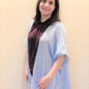 May include: A light blue open-front kimono-style cardigan with short, wide sleeves and a textured fabric finish, layered over a dark maroon sleeveless top and dark blue denim jeans, creating a casual and layered outfit combination.