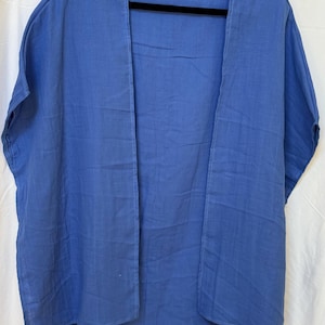May include: A solid blue open-front kimono-style garment with short sleeves and a relaxed fit. The piece features a small white rectangular label at the inner neckline with the text "Mali" printed in black. The fabric has a visible textured weave.