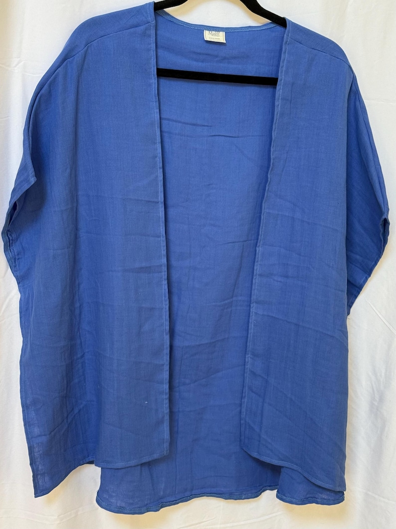 May include: A solid blue open-front kimono-style garment with short sleeves and a relaxed fit. The piece features a small white rectangular label at the inner neckline with the text "Mali" printed in black. The fabric has a visible textured weave.