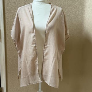 May include: A light beige, open-front kimono-style garment with short, wide sleeves and a textured, semi-sheer fabric. The piece features a simple, loose silhouette with side slits at the hem, designed to drape over the shoulders and torso.