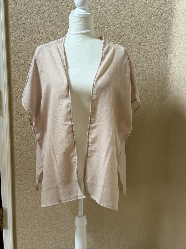 May include: A light beige, open-front kimono-style garment with short, wide sleeves and a textured, semi-sheer fabric. The piece features a simple, loose silhouette with side slits at the hem, designed to drape over the shoulders and torso.