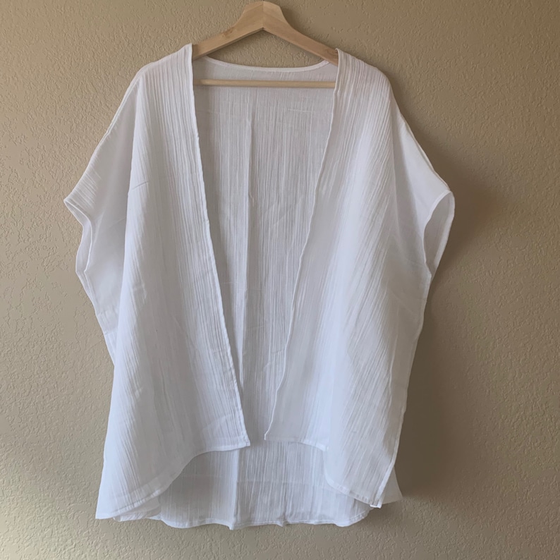 May include: A solid white open-front kimono-style garment featuring a lightweight, crinkled texture. This piece has short, wide sleeves and a loose, flowing silhouette with a slightly longer hemline at the back, hanging on a wooden hanger.