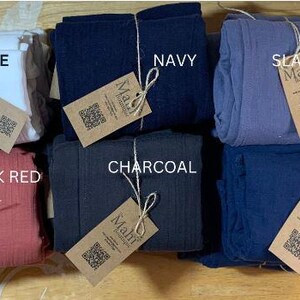 May include: A display of folded fabric bundles in a range of colours: black, white, navy, slate grey, mustard, berry, brick red, charcoal, blue, and ash grey. Each bundle is labelled with a "Mahf" tag.