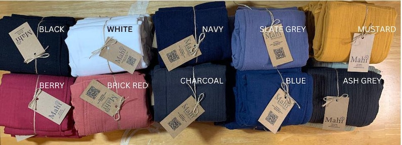 May include: A display of folded fabric bundles in a range of colours: black, white, navy, slate grey, mustard, berry, brick red, charcoal, blue, and ash grey. Each bundle is labelled with a "Mahf" tag.