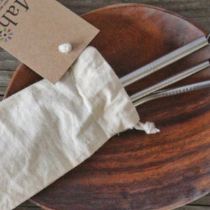 May include: A set of three stainless steel drinking straws in a white cloth bag on a wooden surface. The bag has a tag that says "Lalah" with a colorful flower design.