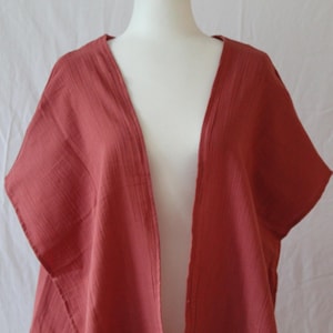 May include: A solid terracotta-colored open-front kimono with short sleeves and a textured, crinkled fabric finish. The garment features a simple, loose-fitting design with a V-neckline, draping naturally over the shoulders and torso.