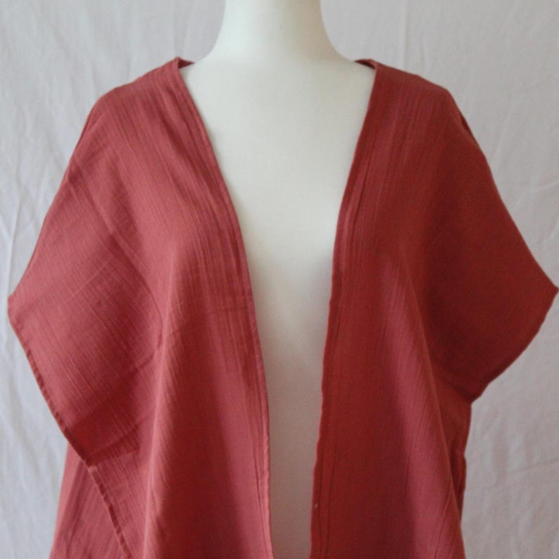 May include: A solid terracotta-colored open-front kimono with short sleeves and a textured, crinkled fabric finish. The garment features a simple, loose-fitting design with a V-neckline, draping naturally over the shoulders and torso.