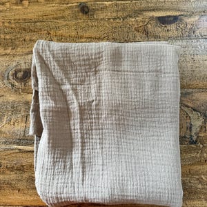 May include: A folded square of light beige muslin fabric featuring a distinct crinkled, multi-layered texture. The textile displays a consistent grid-like weave pattern across its surface, showcasing a soft, matte finish and a neutral earth tone.