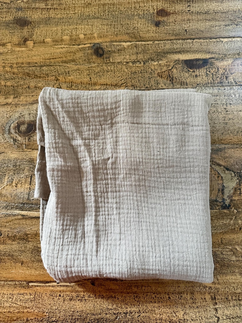 May include: A folded square of light beige muslin fabric featuring a distinct crinkled, multi-layered texture. The textile displays a consistent grid-like weave pattern across its surface, showcasing a soft, matte finish and a neutral earth tone.