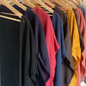 May include: A collection of lightweight, open-front garments with a crinkled texture, each hanging on a wooden hanger. The pieces are displayed in a row, featuring a variety of solid colors including black, navy blue, red, dark brown, mustard yellow, and terracotta.