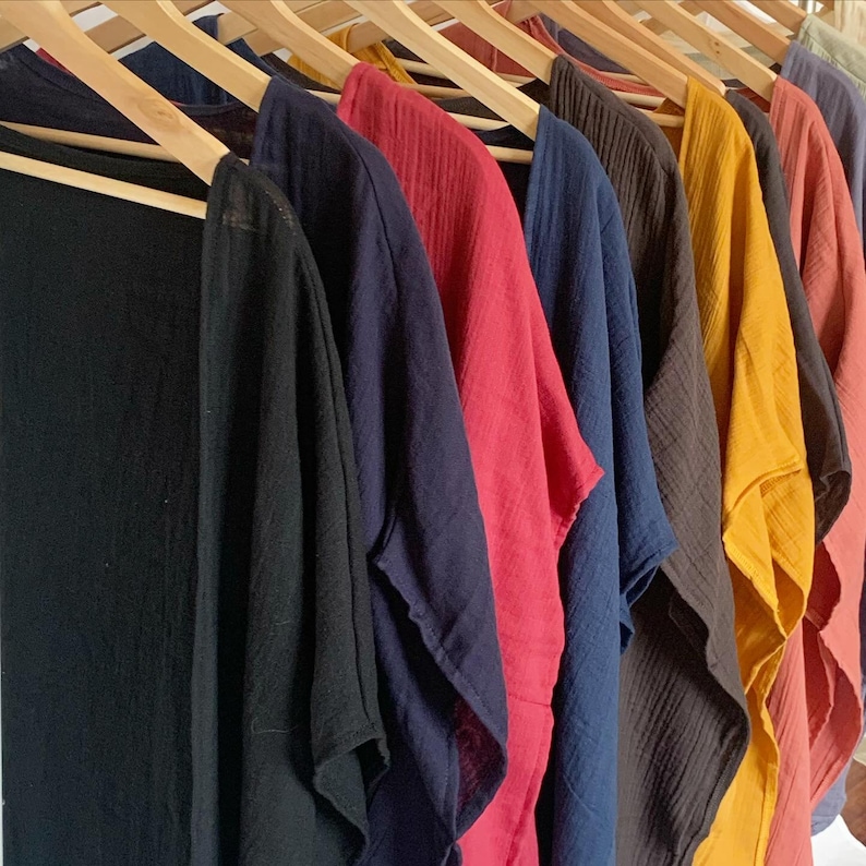 May include: A collection of lightweight, open-front garments with a crinkled texture, each hanging on a wooden hanger. The pieces are displayed in a row, featuring a variety of solid colors including black, navy blue, red, dark brown, mustard yellow, and terracotta.