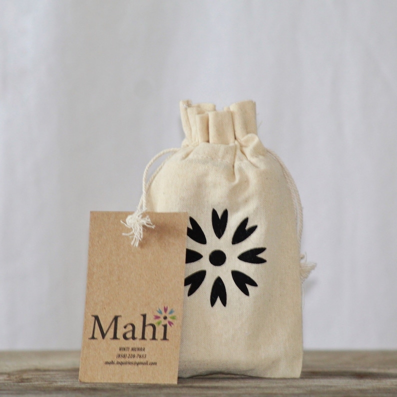 May include: A small, off-white canvas bag with a drawstring closure. The bag features a black floral design printed on the front. A brown paper tag with the text "Mahi" is attached to the bag.