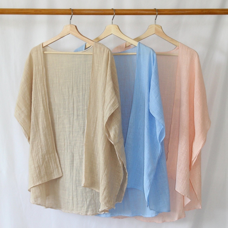 May include: Three open-front kimonos made of lightweight, textured fabric hanging on wooden hangers. The garments are displayed in a row, featuring solid colors of beige, light blue, and soft peach, each with short, wide sleeves and a loose drape.