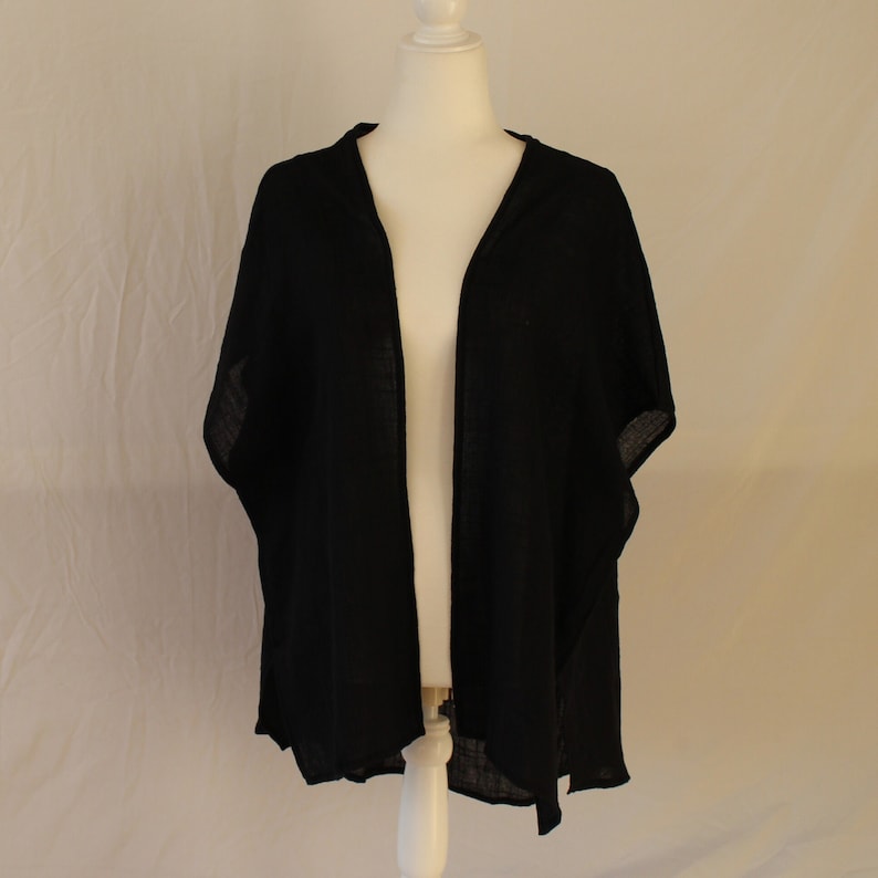 May include: A solid black open-front cardigan with a lightweight, semi-sheer texture and short, wide sleeves. The garment features a simple, draped silhouette with a straight hemline, displayed on a mannequin to show its loose, relaxed fit.