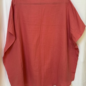 May include: A solid coral-colored tunic top with a round neckline and short, wide sleeves. The garment features a loose, flowy silhouette with a straight hemline and a visible woven fabric texture, hanging on a black hanger against a white background.