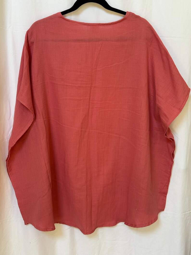 May include: A solid coral-colored tunic top with a round neckline and short, wide sleeves. The garment features a loose, flowy silhouette with a straight hemline and a visible woven fabric texture, hanging on a black hanger against a white background.