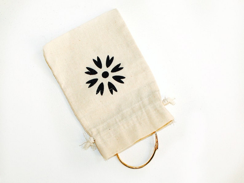 May include: A small, white cotton drawstring bag with a black floral design. The bag is tied with a string and contains a gold bracelet.