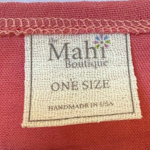 May include: A rectangular white fabric clothing tag sewn onto a textured coral-colored garment. The tag features the text "the Mahi Boutique" with a small colorful flower icon, "ONE SIZE", and "HANDMADE IN USA" printed in black ink.