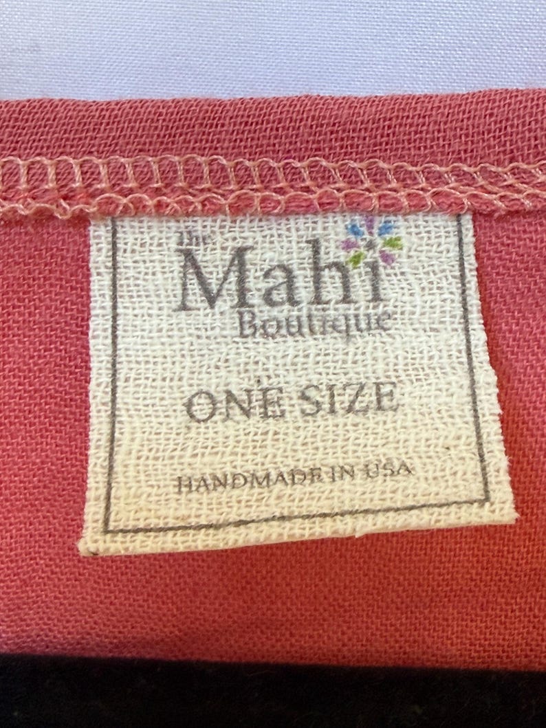 May include: A rectangular white fabric clothing tag sewn onto a textured coral-colored garment. The tag features the text "the Mahi Boutique" with a small colorful flower icon, "ONE SIZE", and "HANDMADE IN USA" printed in black ink.